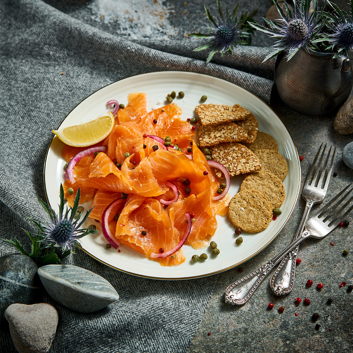 A large orange salmon fillet on a gold rectangular tray, with a white Lochlander smoked salmon label attached. The tray is styled on a gray stone surface with draped gray fabric and scattered river stones.