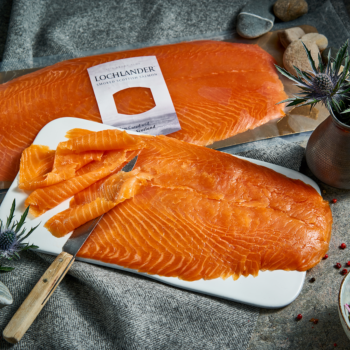 A large orange salmon fillet on a gold rectangular tray, with a white Lochlander smoked salmon label attached. The tray is styled on a gray stone surface with draped gray fabric and scattered river stones.