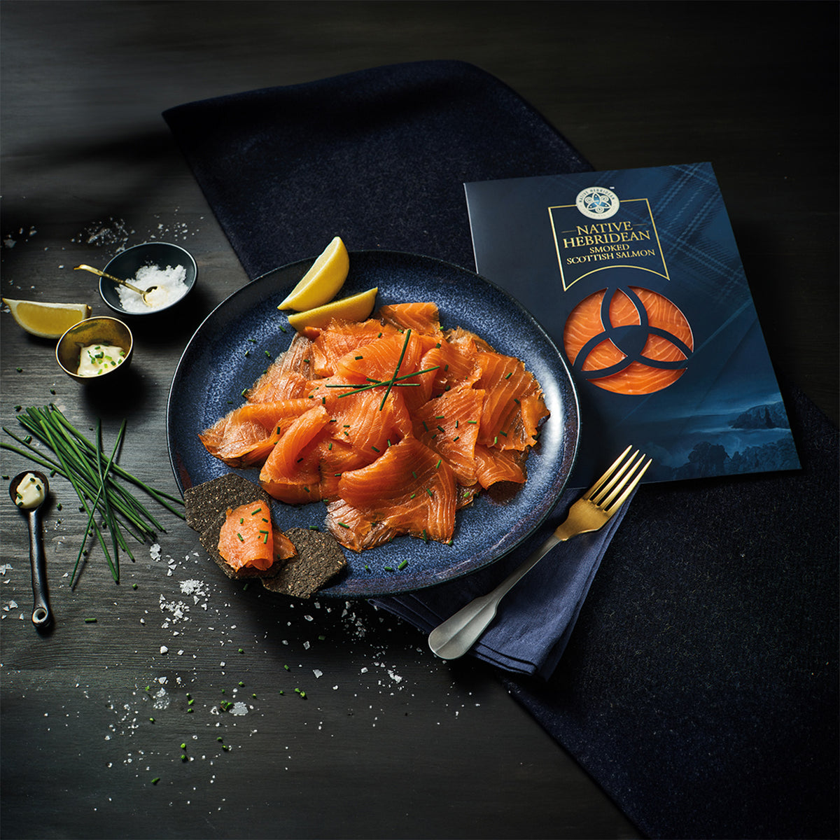 Three packages of Native Herridian Smoked Scottish Salmon are arranged on a dark wooden surface with a black cloth and a small bowl of sea salt.