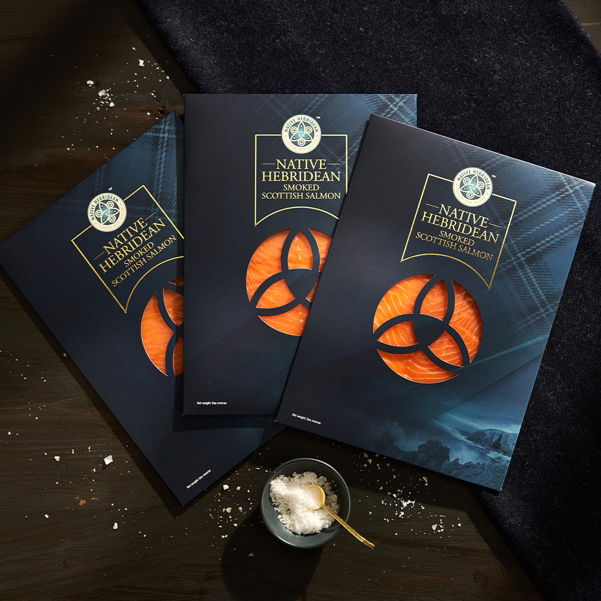 Three packages of Native Herridian Smoked Scottish Salmon are arranged on a dark wooden surface with a black cloth and a small bowl of sea salt.