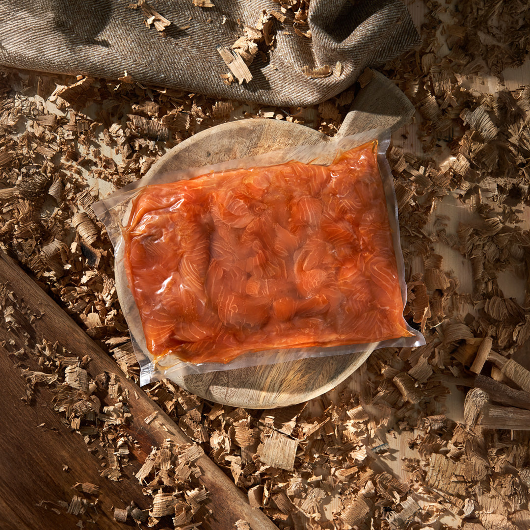 A vacuum-sealed package of orange-red cured meat, likely salami or pepperoni, placed on a round wooden board surrounded by wood shavings and a textured fabric.